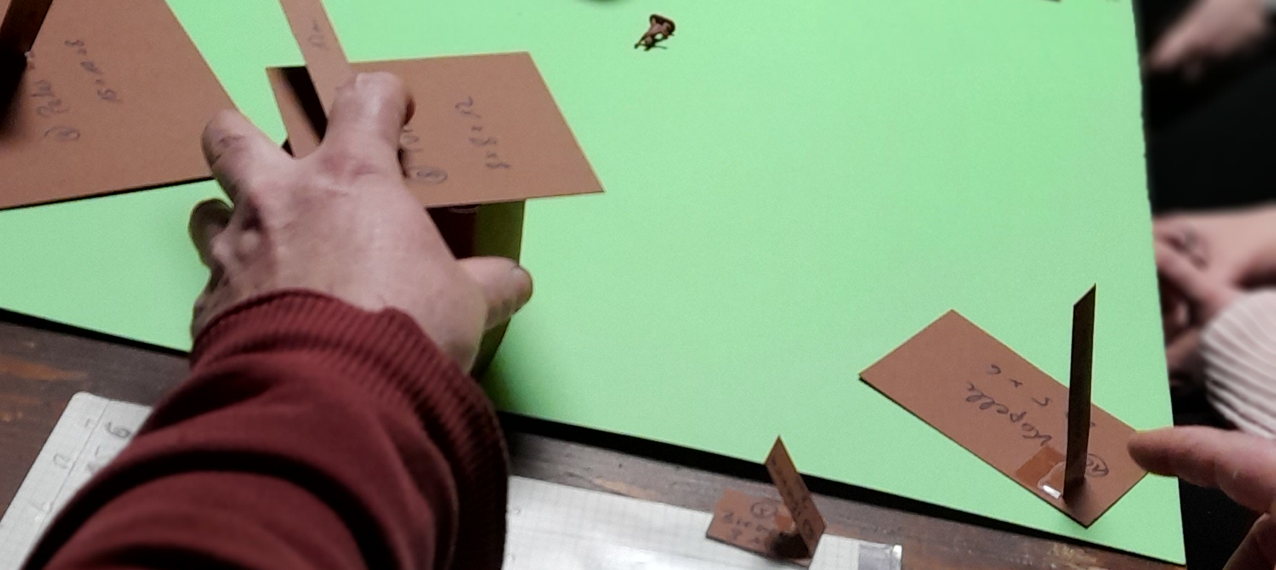 Eine Hand bewegt Modelle aus Pappe auf einer grünen Oberfläche.
